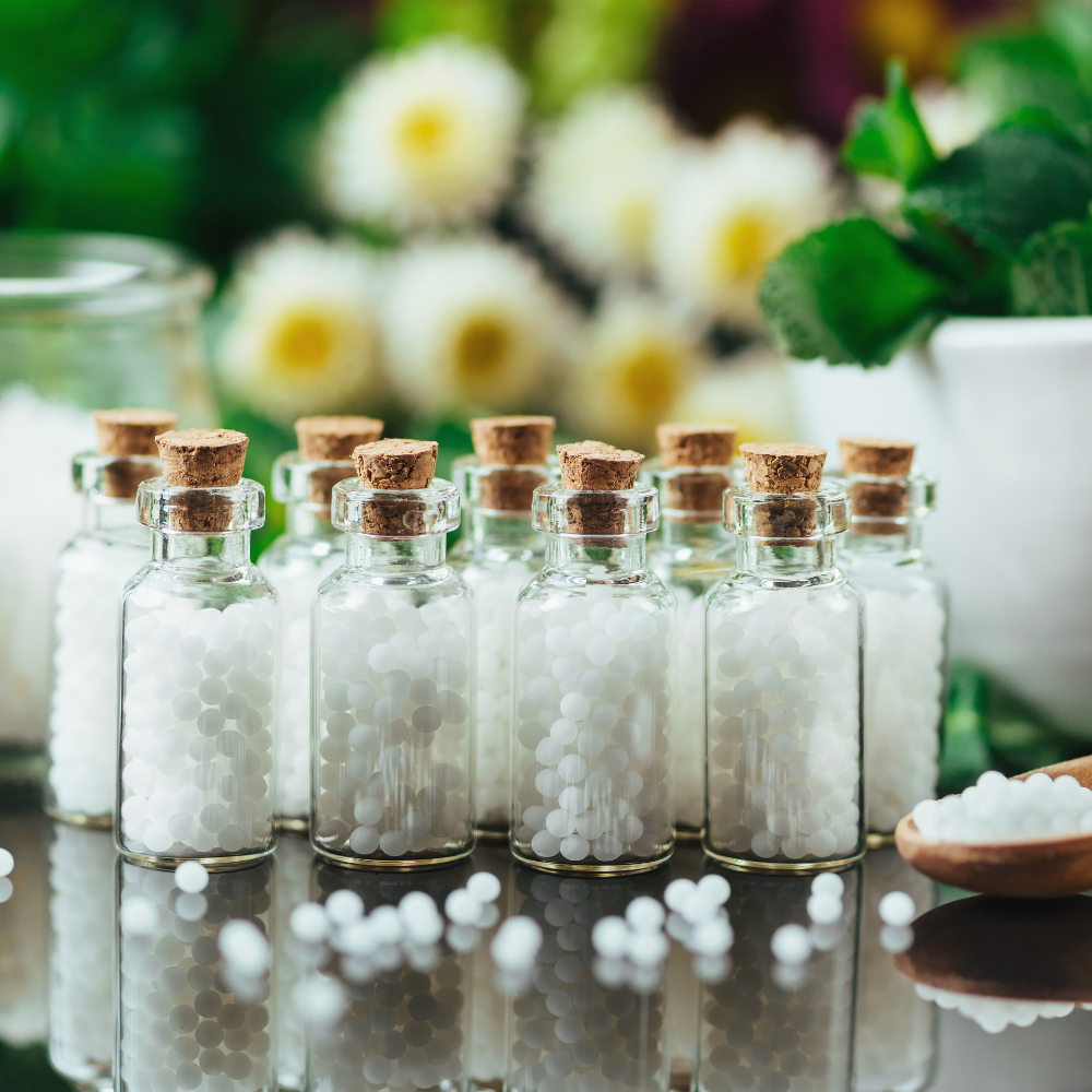Small glass bottles with cork stoppers,  containing homeopathic remedies, arranged on a surface with a blurred background featuring flowers and a cup.